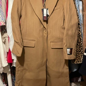 Camel kappa från Tommy Hilfiger - Snygg camel-färgad kappa från Tommy Hilfiger i storlek 36, känns mer som 38, jag upplever den som stor i storleken. jag upplever den som stor i storlek, därför säljs den! Ny med etikett, ord pris 6000kr. Jag har kvitto.   (ber om ursäkt att den är skrynklig, legat i kartong)