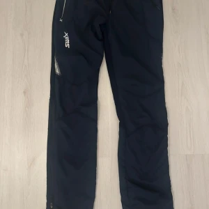 Svarta täckbyxor från Swix herr S - Svarta täckbyxor från Swix i herrstorlek S. Byxorna har elastisk midja med snörning, dragkedjor vid bensluten och meshdetaljer på sidorna. Perfekta för vinterns utomhusaktiviteter och träning. Lätta och smidiga med sportig look.