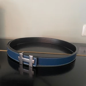 blått skärp med H-spänne - Snyggt blått skärp med ikoniskt H-spänne i silverfärgad metall. Bältet är gjort i äkta skinn med vita kontrastsömmar och svart insida. Perfekt accessoar för att lyfta din outfit med en lyxig touch.