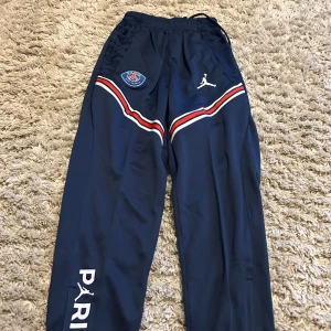 PSG x Jordan blå träningsbyxor - Mörkblå träningsbyxor från PSG x Jordan med vita och röda detaljer, broderad PSG-logga och Jumpman på framsidan. Byxorna har elastisk midja med snörning och tryckt text på benet. Perfekta för fotboll eller chill.