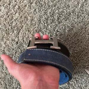 H3rmès vändbart skärp i skinn och jeans - Snyggt vändbart H3rmès-bälte med klassiskt H-spänne i silver. Ena sidan är svart skinn med vit söm, andra sidan är blå jeans med kontrastsöm. Perfekt accessoar för att lyfta din outfit med en lyxig touch och cool detalj.