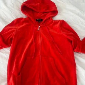 Juicy Couture röd velour hoodie - Juicy Couture hoodie i röd velour med dragkedja framtill, huva med snörning och känguruficka. Långärmad och supermjuk, perfekt för en chill och trendig look. Klassisk logga på insidan av kragen. Använd 2 gånger.