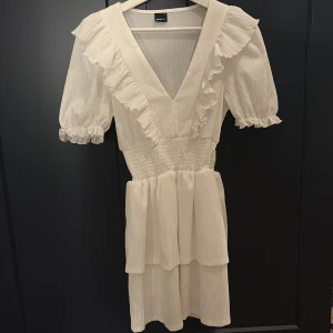 Vit volangklänning från Gina Tricot - Superfin vit kortklänning från Gina Tricot med v-ringning, volangdetaljer och puffärmar. Klänningen har smock i midjan och dubbla lager i kjolen för extra volym. Perfekt för sommarens alla tillfällen!