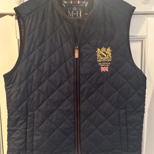 Quiltad blå väst från Morris - Snygg quiltad väst från Morris i mörkblått med guldfärgat broderat emblem och brittisk flagga på bröstet. Västen har dragkedja framtill, två fickor och klassisk krage. Perfekt för lager-på-lager och stilren look.