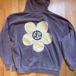 brunt hoodie med blomma och peace-tryck - brunt hoodie från Urban Outfitters BDG med stor beige blomma och texten 'PEACE' på ryggen. Framsidan har en mindre blomma och peace-symbol. Hoodien har känguruficka, huva och är långärmad. Perfekt för dig som gillar retro vibes och coola tryck.
