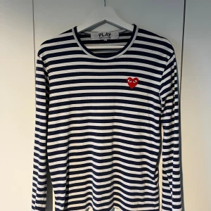 Randig långärmad tröja Comme des Garçons - Säljer en långärmad tröja från Comme des Garçons Play i marinblått och vitt med klassiskt randigt mönster. Tröjan har ett rött hjärta med ögon broderat på bröstet och är gjord i mjuk bomull. Perfekt för dig som gillar stilrena och ikoniska plagg. Storlek M sitter som S