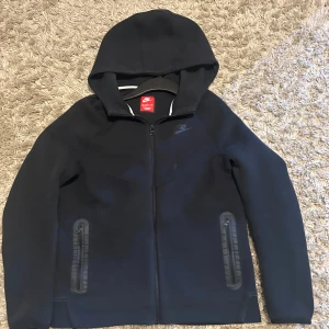 Svart Nike hoodiejacka XL - Svart hoodiejacka från Nike i storlek XL. Jackan har dragkedja framtill, två fickor med dragkedja och en stilren look. Perfekt för dig som gillar sportig och clean stil. Mjuk och bekväm med huva för extra chill-vibe.