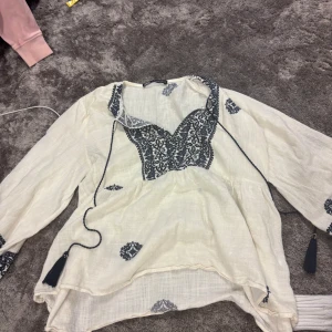 Vit blus med broderi och knytband - Vit blus i luftig bomull med mörkblå broderade detaljer och knytband med tofsar vid halsen. Blusen är i fint skick korrekt till storlek. Man fryser ej i den💕