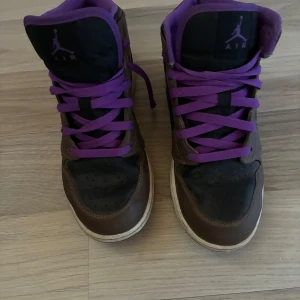Nike Air Jordan 1 High brun/lila - Snygga Nike Air Jordan 1 High sneakers i brunt och svart skinn med lila detaljer och lila snörning. Klassisk Jordan-logga på sidan och plös. Hög modell med vit sula och lila innerfoder. Perfekta för dig som vill sticka ut med färg och stil.