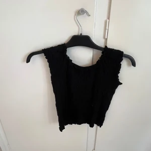 Svart croppad ärmlös topp - Säljer en svart croppad topp med ärmlös design och rynkat tyg. Toppen har en lätt vågig kant nertill och vid axlarna, vilket ger en trendig look. Perfekt att matcha med höga jeans eller kjol för en snygg outfit. Stretchig så passar någon storlek större