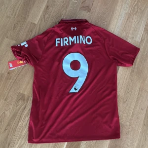 Liverpool Firmino #9 matchtröja M - Helt ny Liverpool FC matchtröja från New Balance i vinröd färg med vita detaljer. Tröjan har korta ärmar, krage och tryck med Standard Chartered, LFC-logga samt Firmino och nummer 9 på ryggen. Tillverkad i lätt funktionsmaterial som andas. Ni är alltid välkomna att skriva om ni har frågor 😄