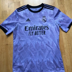Real Madrid lila bortatröja Adidas - Säljer en lila Real Madrid bortatröja från Adidas med svart krage och svarta detaljer. Tröjan har geometriskt mönster, korta ärmar och är tillverkad i lätt och ventilerande Aeroready-material. Perfekt för dig som vill sticka ut på planen eller läktaren.