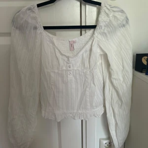 Vit blus - Supersöt vit blus, långa puffärmar och smockad överdel. Blusen har små knappar framtill, volangkant nertill och en luftig, romantisk känsla. Perfekt för dig som gillar en feminin och trendig stil.