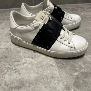 Valentino Rockstud vita sneakers - Snygga vita Valentino Rockstud sneakers med svart läderdetalj och ikoniska nitar. Skorna har snörning, rund tå och platt sula. Tillverkade i skinn med coola pyramidnitar runt hälen och svart band över vristen. Perfekta för dig som vill sticka ut med en lyxig streetstyle. Nypris 7000