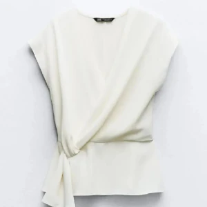 Vit omlottopp från Zara - Stilren vit omlottopp från Zara med knyt i sidan. Toppen har v-ringning och är ärmlös, vilket ger en fräsch och modern look. Perfekt att matcha med jeans eller kjol för en clean stil. Säljer då den inte kommit till användning!