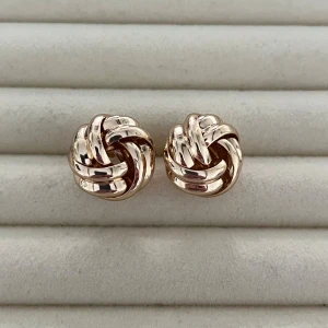 Guldiga knutörhängen i metall - Snygga örhängen i guldig metall med knutdesign. De är blanka och har en rund, flätad form som ger en cool och tidlös look. Perfekta för att lyfta vilken outfit som helst med lite extra shine.