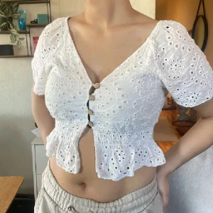 Vit broderad blus med knappar - Supersöt vit blus med broderade hålmönster, korta puffärmar och v-ringning. Toppen är croppad med smockad midja och volangkant nertill. Träknappar framtill ger en extra detalj. Perfekt för sommarens alla tillfällen!