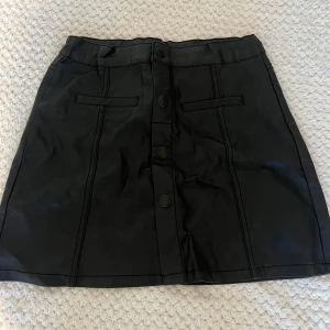 Svart skinn kjol - Svart kort kjol från Lindex i skinnimitation med snygga sömdetaljer och knappar framtill. Kjolen har två dekorativa fickor framtill och en rak passform som ger en cool vibe. Perfekt för dig som gillar en stilren och edgy look.pris kan diskuteras och den passar xs och s🤍