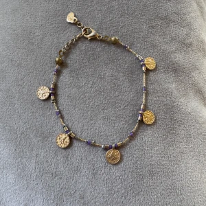 Guldigt armband med lila pärlor - Snyggt armband med små guldmynt och lila pärlor. Kedjan är tunn och har justerbar längd med ett hjärtformat lås. Perfekt accessoar för att ge din outfit lite extra bling och färg.