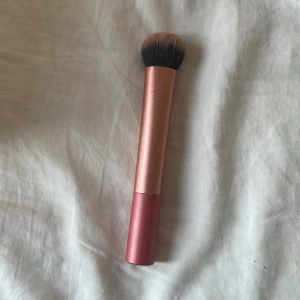 Sminkborste från Real Techniques - Säljer en sminkborste från Real Techniques med roséguldfärgat handtag och täta, mjuka borst i svart och beige. Perfekt för applicering av foundation eller puder. Handtaget är i metall och borsten är syntetisk. Sminkborsten tvättas såklart innan!