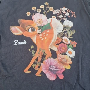 Svart Bambi linne från Disney - Svart ärmlös t-shirt/linne från Disney med stort färgglatt tryck av Bambi och blommor på framsidan. Snygg rund halsringning och mjukt bomullsmaterial. Storlek M, passar S-L beroende på hur man vill ha passformen.