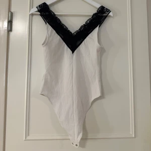 Vit body med svart spets från Zara - Supersnygg vit body från Zara med djup v-ringning och bred svart spetskant. Ärmlös modell med knäppning i grenen och lätt ribbad struktur i tyget. Perfekt för dig som vill ha en stilren men ändå detaljrik topp. Säljer för att den inte kommer till användning 