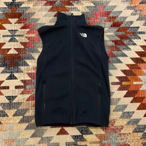 Mörkblå fleeceväst från The North Face XS - Svart fleeceväst från The North Face i storlek XS. Västen har hög krage, hel dragkedja framtill och två fickor med dragkedja. Broderad logga på bröstet. Perfekt lager-på-lager-plagg för kyliga dagar.