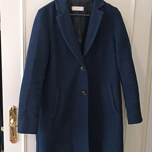 Mörkblå ullkappa från Féraud Paris - Snygg mörkblå kappa från Féraud Paris i klassisk rak modell med två knappar framtill och diskreta fickor. Tillverkad i ull, perfekt för kyliga dagar. Stilsäker och tidlös design som lyfter varje outfit.