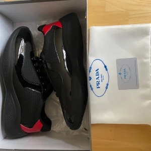 Svarta sneakers från Prada med röda detaljer - Snygga svarta sneakers från Prada med röda detaljer på hälen och logga på plösen. Skorna har en mix av blankt skinn och mesh, samt svarta snören och en robust svart sula. Perfekta för dig som vill ha en exklusiv och modern look.