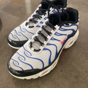 Nike Air Max Plus Tn sneakers blå/vit - .Nike Air Max Plus Tn sneakers med vit mesh, blå vågiga detaljer och svarta partier. Swoosh-logga i rött på sidan och Tn Air-märke bak. Sulan är vit med röda och gula inslag undertill. Skorna har svart snörning och en sportig vibe