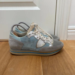 Phillipe model skor - Säljer ett par snygga sneakers från Philippe Model i grått och ljusblått med inslag av vitt. Skorna har ovandel i mocka och mesh, vita snören och en platt sula med blå och beige ränder. Perfekta för dig som gillar stilrena och sportiga skor. Skorna är i storlek 43 och passar perfekt till sommaren med ett nypris på cirka 3000 kr!
