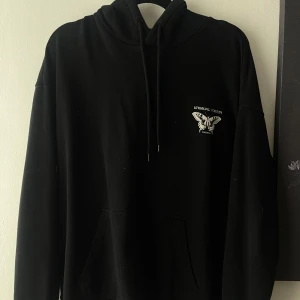 Svart hoodie med fjärilstryck Unusual Vision - Svart hoodie med vit fjäril och botaniskt tryck både fram och bak. Perfekt för dig som gillar streetstyle och unika prints.