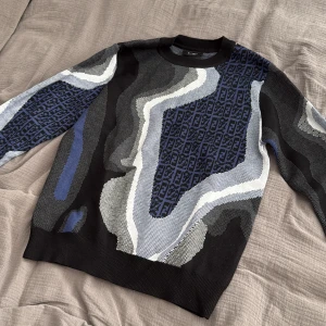 Unik Fendi tröja - Snygg stickad tröja från Fendi med abstrakt mönster i svart, blått, grått och vitt. Tröjan har rund halsringning och långa ärmar. Fendi-loggan är invävd i det blå partiet på bröstet och ärmarna. Perfekt för dig som gillar statement-plagg.