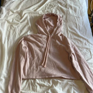 Ljusrosa croppad hoodie med huva - Säljer en ljusrosa croppad hoodie med långa ärmar och ribbade muddar. Tröjan har en klassisk huva med snörning och är tillverkad i mjuk bomullsmix. Perfekt för en chill och avslappnad stil.
