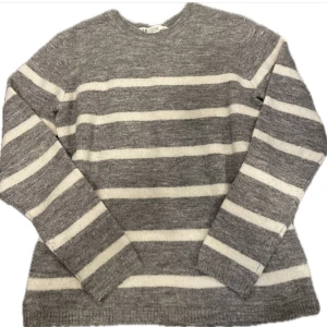 Grå randig stickad tröja - Stickad tröja i grått och vitt med breda horisontella ränder, rund halsringning och långa ärmar. Perfekt i sommar lite kyligare dagar både till kjol eller jeans. Står storlek 134/140 men passar XXS/XS.