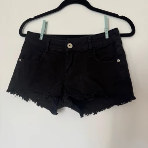 Svarta jeansshorts med fransig kant - Svarta jeansshorts med rå, fransig kant och klassisk femficksmodell. Shortsen har låg midja och silverfärgade nitar vid fickorna. Perfekta för en chill sommarlook med lite edge.