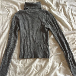 Grå ribbad polotröja - Snygg grå ribbad polotröja med lång ärm och tight passform. Perfekt för lager-på-lager och enkel att styla till olika outfits. Polokragen är hög och ger en clean look. Materialet är stretchigt och mjukt, vilket gör den superbekväm.