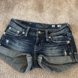 Miss Me jeansshorts med detaljer - Snygga jeansshorts från Miss Me i mörkblå denim med slitningar och uppvikta benslut. Fickorna bak har blingiga knappar och broderade detaljer. Klassisk femficksmodell med låg midja och coola kontrastsömmar. Perfekta för sommaren!