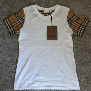 Vit Burberry t-shirt med rutiga ärmar - Snygg vit t-shirt från Burberry med klassiska beige-rutiga ärmar i deras ikoniska mönster. T-shirten har rund halsringning och är i mjuk bomull. Perfekt för dig som vill ha en stilren look med en touch av lyx.