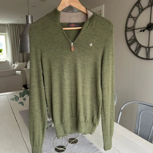 Olivgrön half zip-tröja Morris - Olivgrön långärmad tröja från Morris med half zip och liten broderad logga på bröstet. Tröjan är stickad i 100% merinoull och perfekt för lager-på-lager och snygg till jeans. Tröjan är sydd i ena armhålan en gång men detta syns knappt.