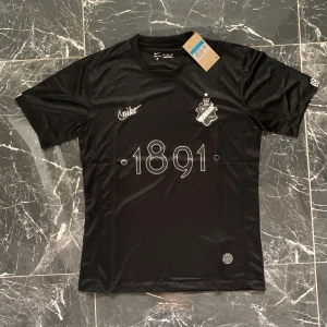Svart AIK fotbollströja Nike M - Snygg svart AIK fotbollströja från Nike med klubbmärke på bröstet, trycket '1891' framtill och texten 'SMOKINGLIRARE' i nacken. Tröjan är kortärmad och tillverkad i ett lätt, sportigt material som andas. Perfekt för dig som vill visa ditt AIK-stöd på matchen eller träningen.