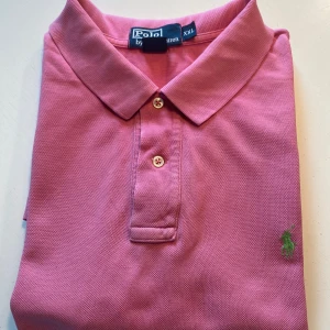 Rosa pikétröja från Polo Ralph Lauren - Snygg rosa pikétröja från Polo Ralph Lauren i klassisk modell med krage och två knappar. Tröjan har det ikoniska gröna broderade logot på bröstet och är gjord i bomull. Perfekt för en chill och stilren look.