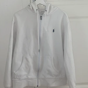 Vit hoodie från Polo Ralph Lauren - Vit hoodie från Polo Ralph Lauren med dragkedja och huva. Klassisk design med broderad logga på bröstet och känguruficka framtill. Tillverkad i mjuk bomull, perfekt för chill dagar. Snygg och enkel att matcha med allt. Passar för Storlek M och storlek S
