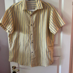 Gul- och blårandig kortärmad skjorta - Kortärmad skjorta med gula och blå ränder, klassisk krage och knäppning framtill. Skjortan har en avslappnad passform och är perfekt för varma dagar. Tillverkad i mjukt bomullstyg som känns skönt mot huden.