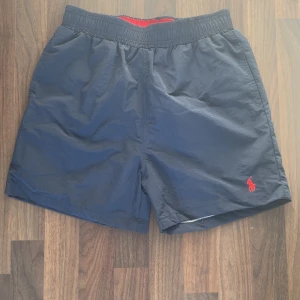Mörkblå badshorts från Polo Ralph Lauren - Snygga mörkblå badshorts från Polo Ralph Lauren med röd broderad logga framtill. Klassisk modell med elastisk midja och innerbyxa i mesh. Perfekta för stranden eller poolen, med en bakficka och sportig vibe.
