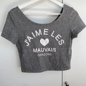 Grå croppad topp med tryck - Grå croppad topp med korta ärmar och vitt tryck framtill där det står 'J'aime les mauvais garçons' och ett hjärta. T-shirten har en rundad, bred urringning och är tillverkad i mjukt bomullsmaterial. Perfekt för en chill och trendig look.