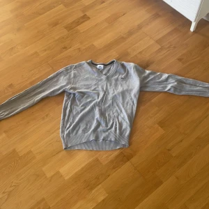 Grå stickad Lacoste tröja - Grå stickad tröja från Lacoste med klassisk v-ringning och diskret krokodillogga på bröstet. Långärmad modell med ribbade muddar och nederkant. Perfekt för dig som gillar stilren och enkel design.