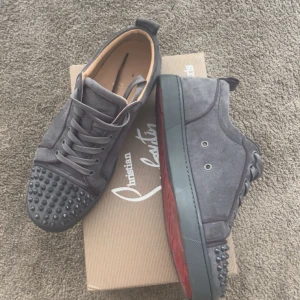 Grå mocka sneakers från Christian Louboutin - Snygga grå sneakers i mocka från Christian Louboutin med ikonisk röd sula och coola nitar på tån. Skorna har snörning och en rund tå, perfekt för dig som vill sticka ut med en lyxig streetstyle-look.