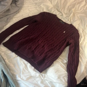 Vinröd Ralph Lauren tröja - Säljer en vinröd kabelstickad tröja från Polo Ralph Lauren. Tröjan har lång ärm, rund halsringning och den klassiska broderade loggan på bröstet. Perfekt för dig som gillar preppy stil och vill ha något snyggt och bekvämt till hösten och vintern.