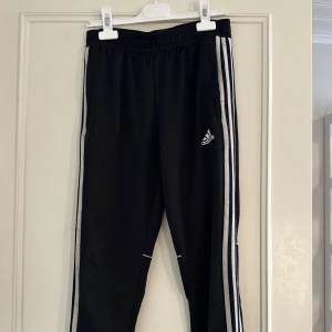 Svarta Adidas träningsbyxor med dragkedja - Svarta träningsbyxor från Adidas med klassiska vita ränder längs sidorna och Adidas-logga på låret. Byxorna har knappt används men säljer pågrund av att de inte passar mig längre.